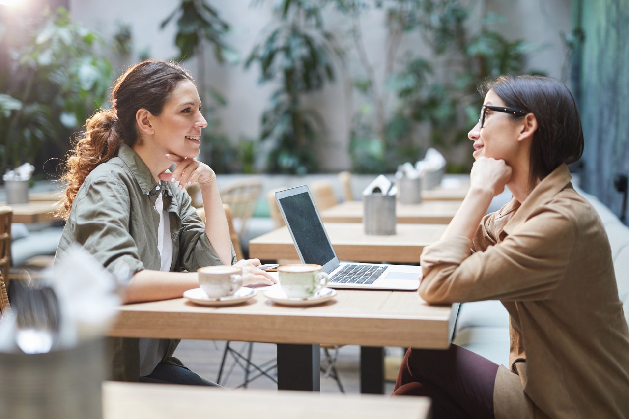 Zwei Frauen sitzen sich in einem hellen, mit Pflanzen dekorierten Café an einem Tisch mit zwei Kaffeetassen und einem Laptop gegenüber und unterhalten sich. © Seventyfour / stock.adobe.com