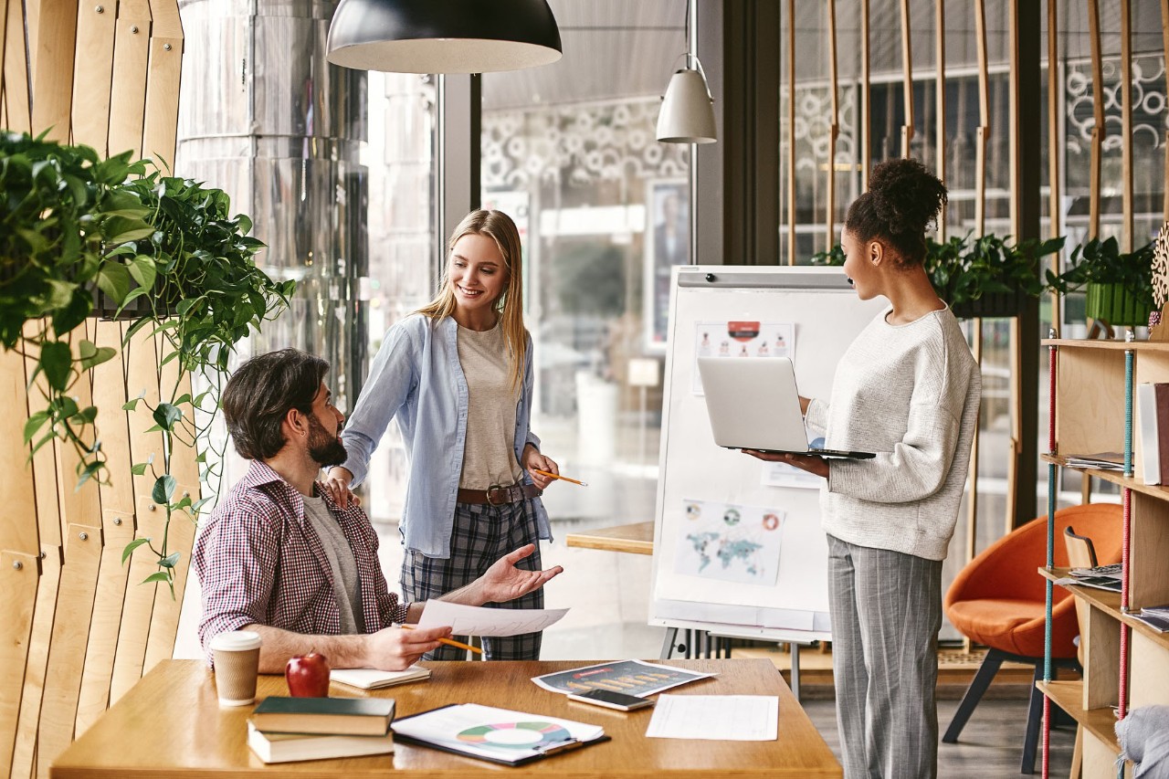 Drei Personen, ein Mann und zwei Frauen, in Meeting-Situation mit Laptop und Flipchart in einem hellen, modernen Büro. © Friends stock / stock.adobe.com