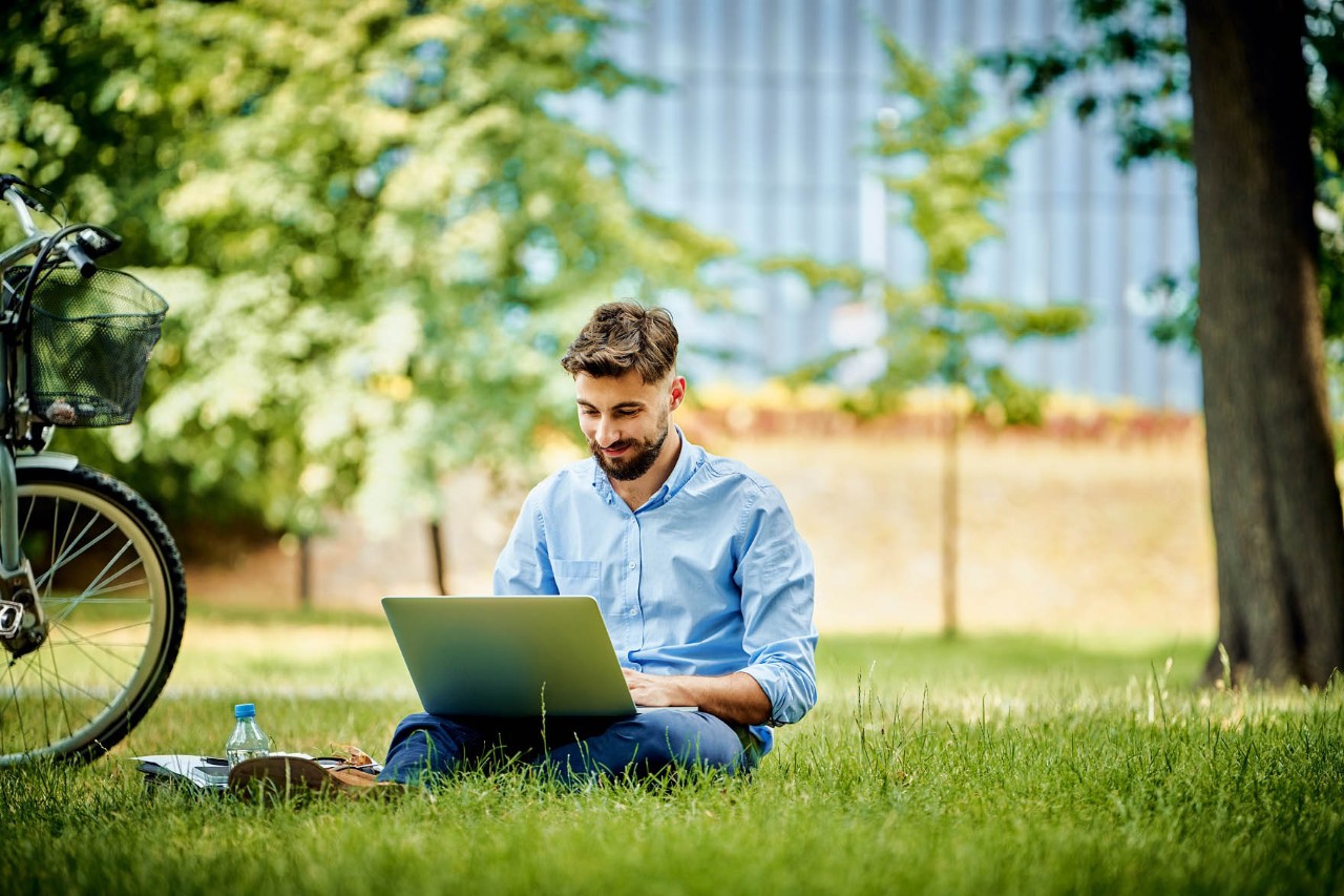 Ein junger Mann sitzt in parkartiger Umgebung bei gutem Wetter im Gras und arbeitet lächelnd an seinem Laptop. © baranq / stock.adobe.com