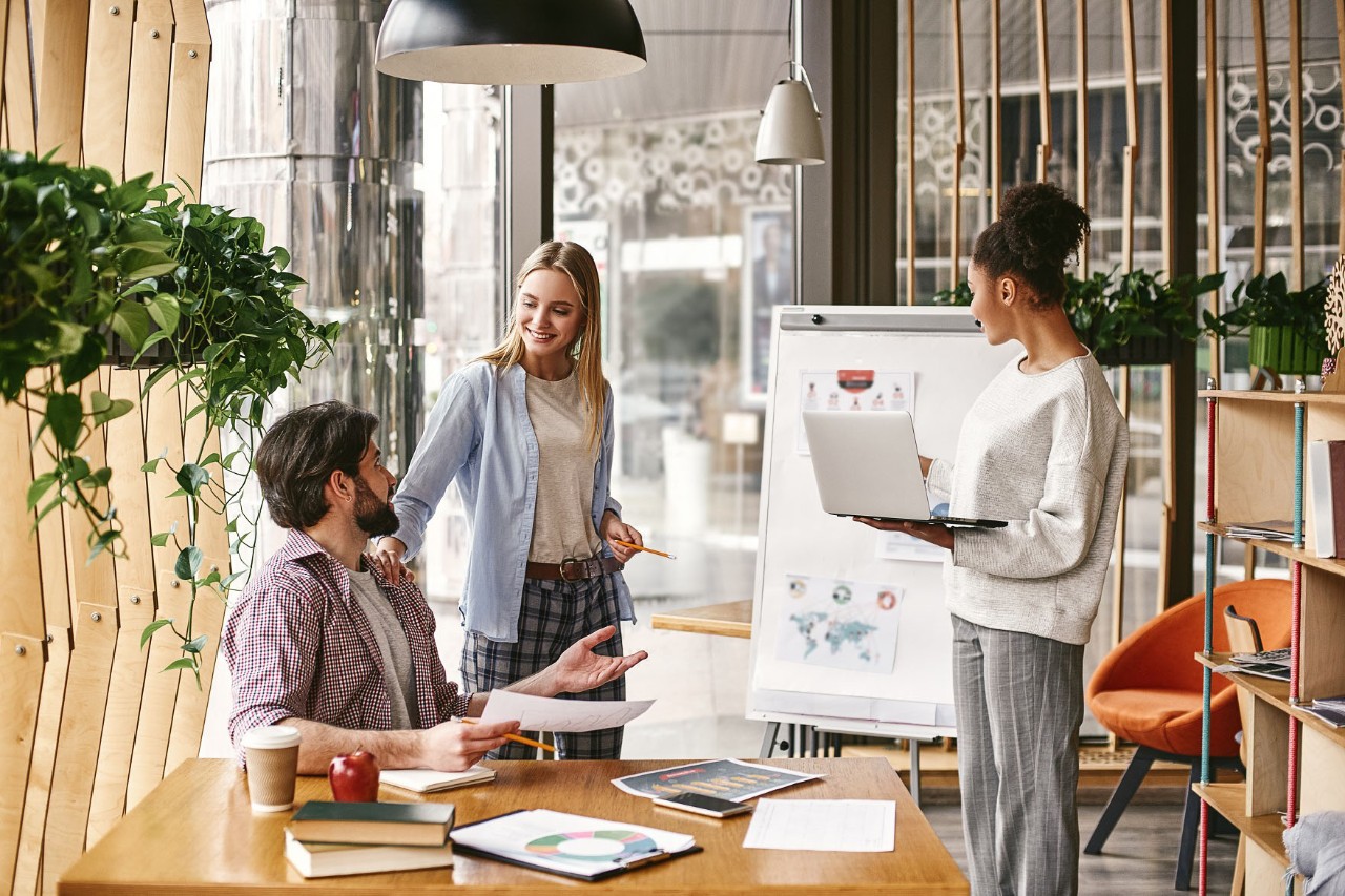 Three people, a man and two women, in a meeting situation with a laptop and a flipchart in a bright, modern office. © Friends stock / stock.adobe.com